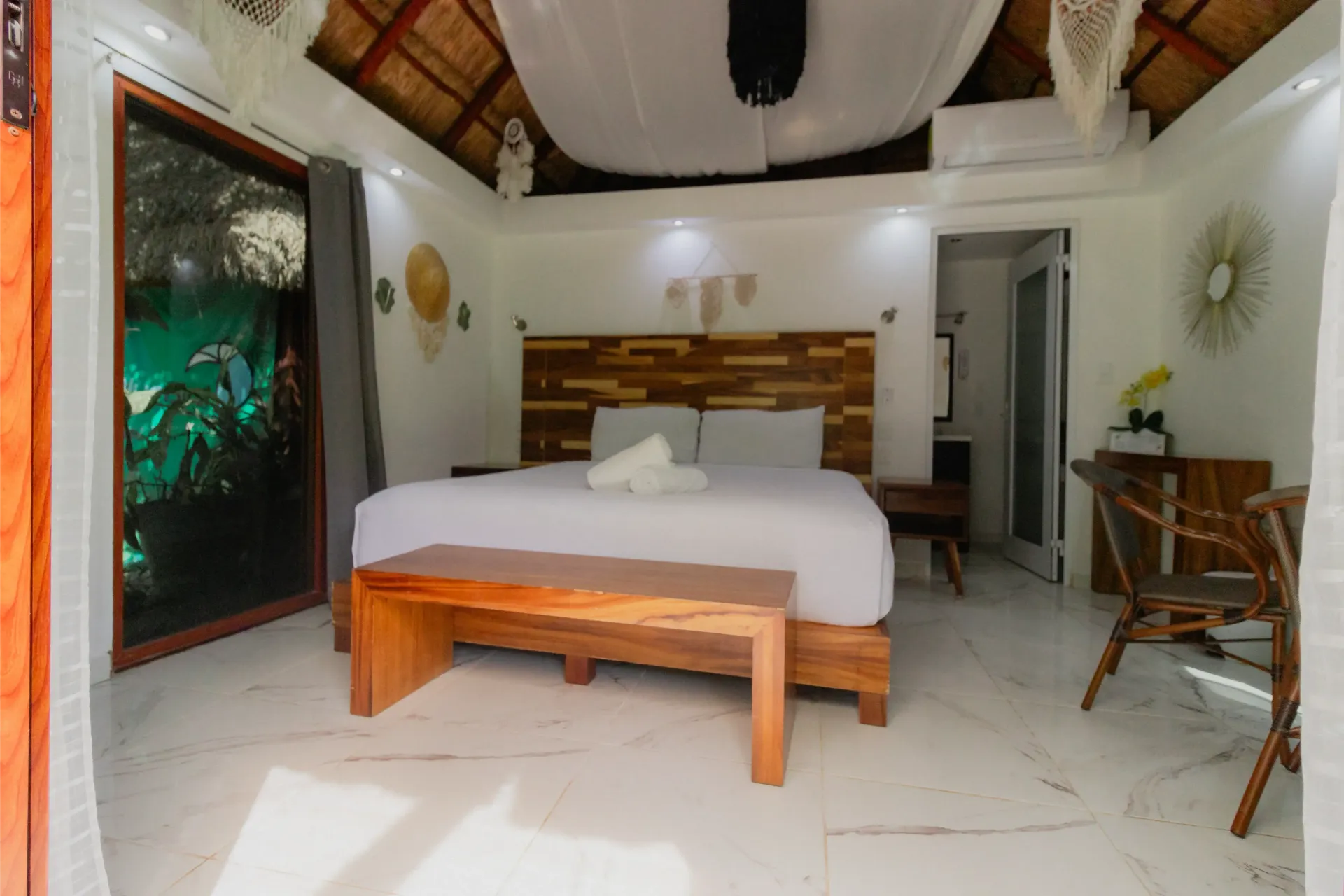 Habitación luminosa con una cama grande, banco de madera, paredes blancas y puerta que da a un balcón con vegetación.