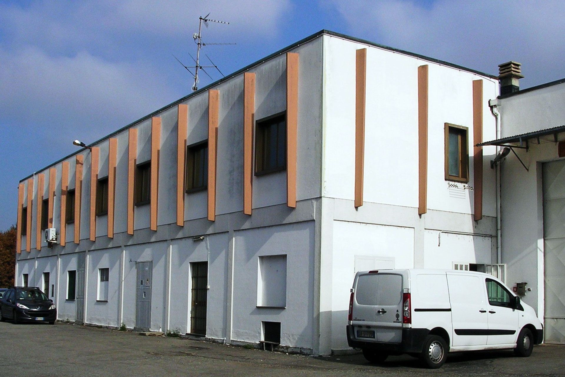 Edificio bianco a due piani con accenti verticali arancioni, piccole finestre e un furgone parcheggiato fuori.