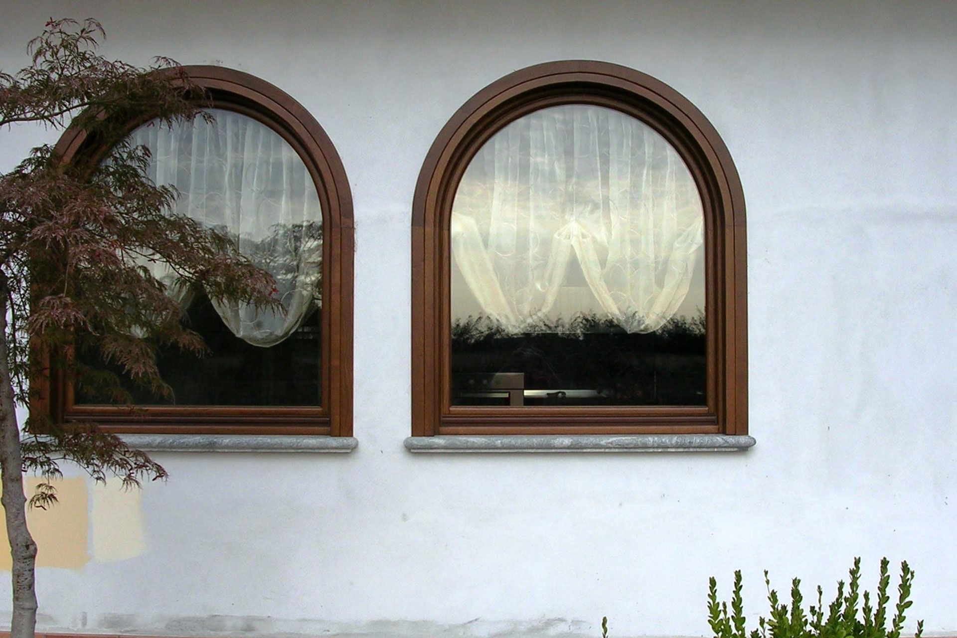 Due finestre ad arco con cornice marrone su una parete bianca con tende trasparenti che riflettono l'esterno.