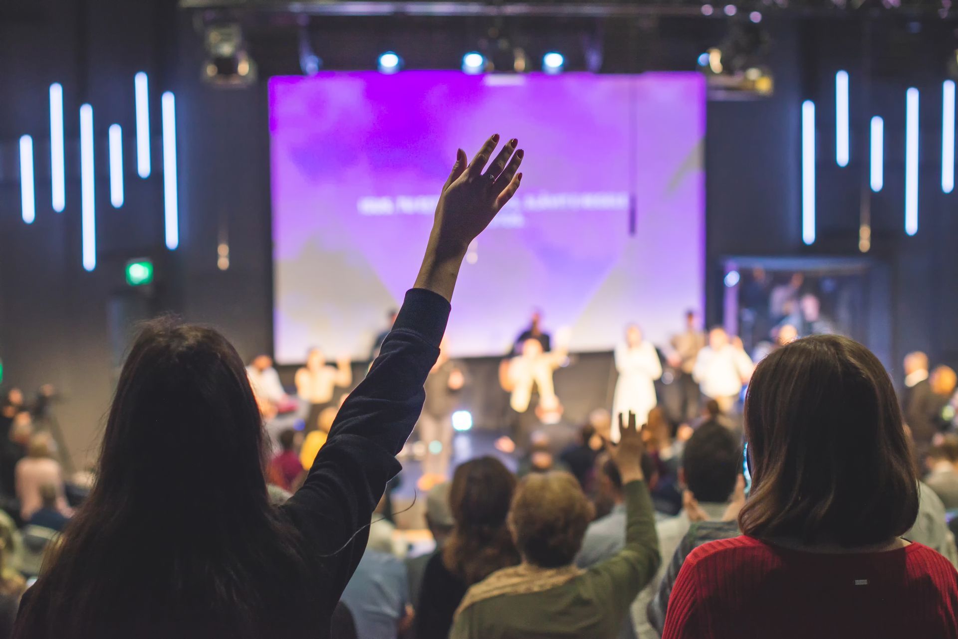 People with raised hands in a church service with a stage and screen in the background.