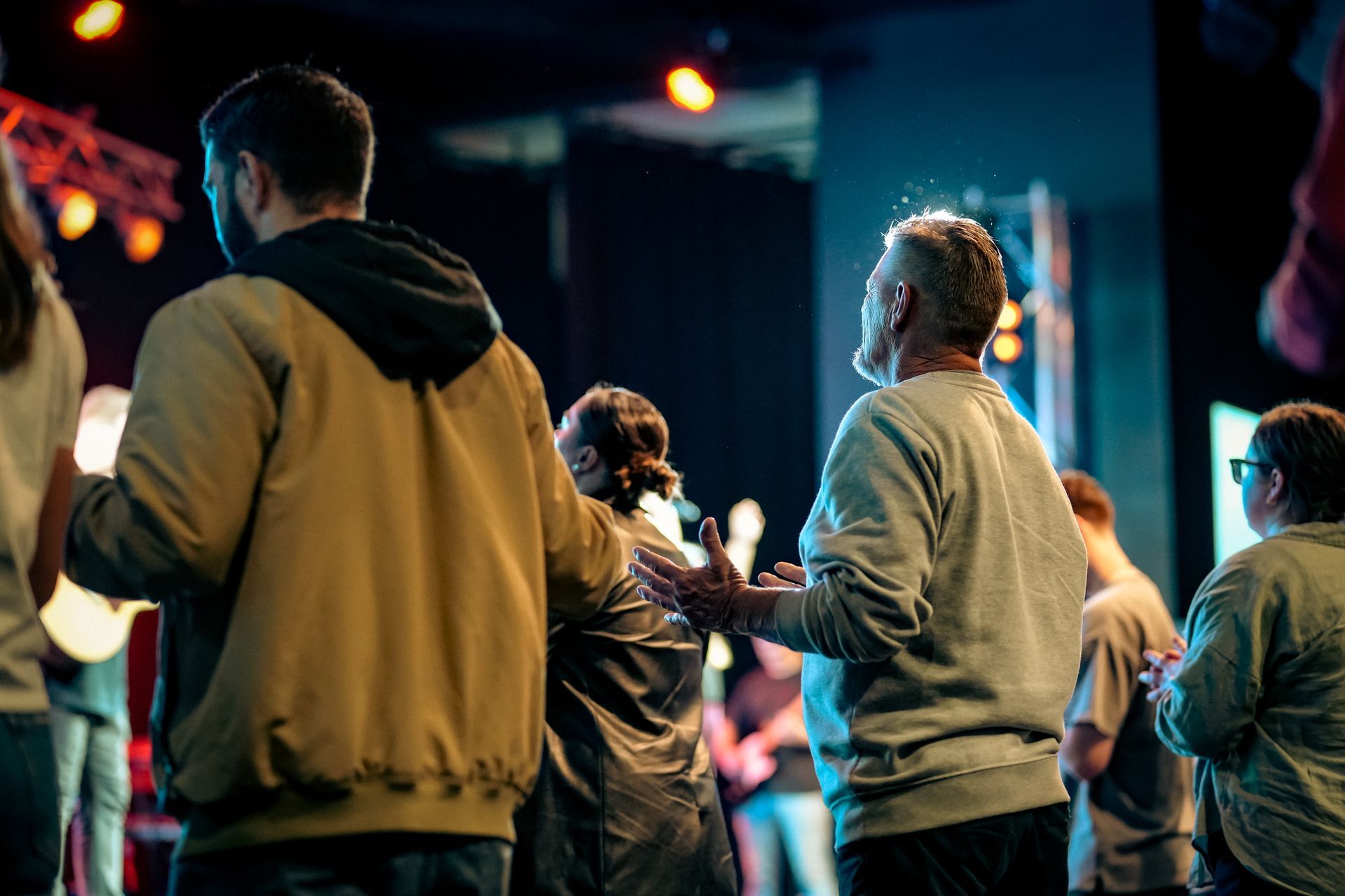 People with hands raised, in a room with stage lighting. Some in casual clothing, possibly at a concert or performance.