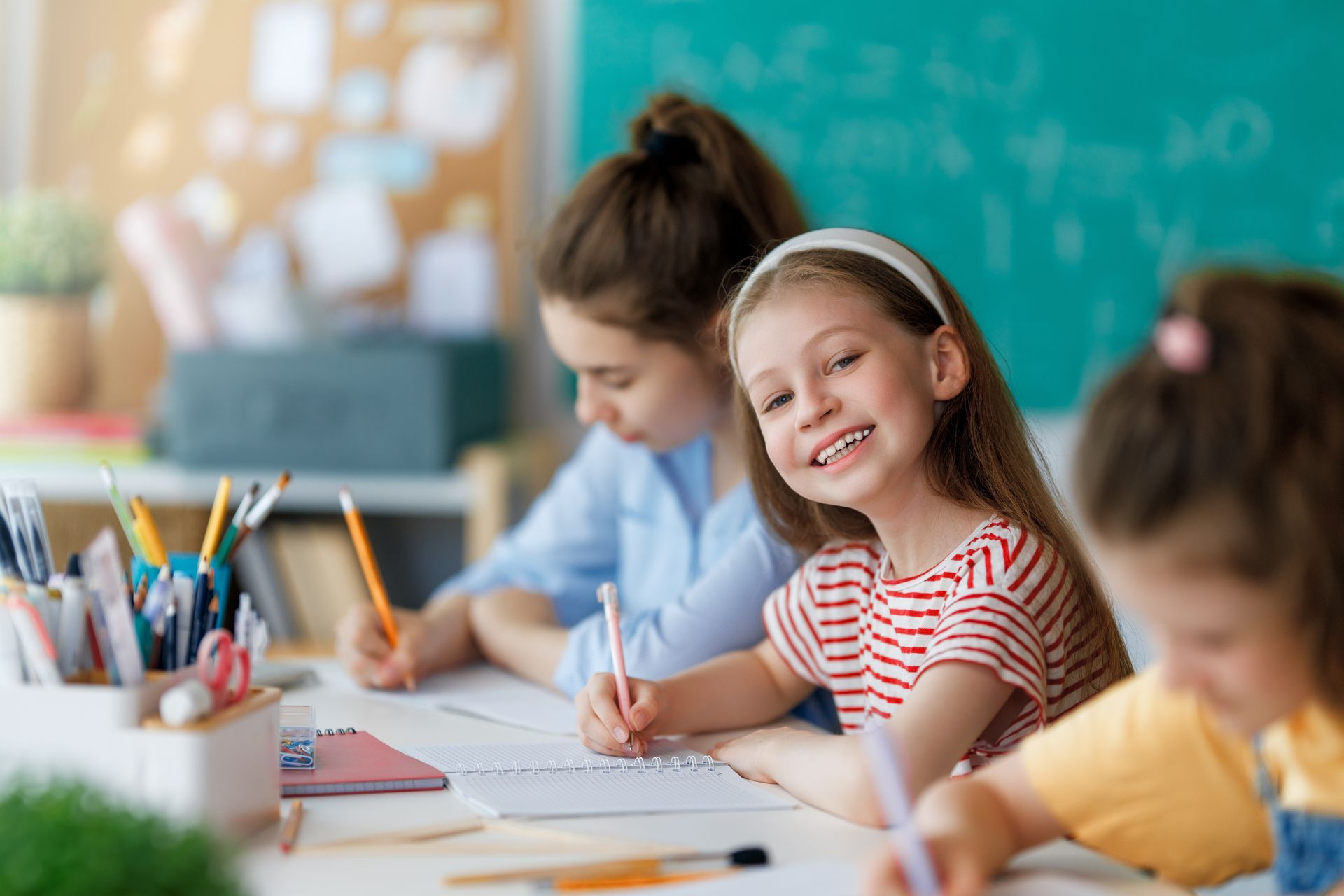 Children at desks in a classroom, writing with pencils. One girl smiles at the camera.