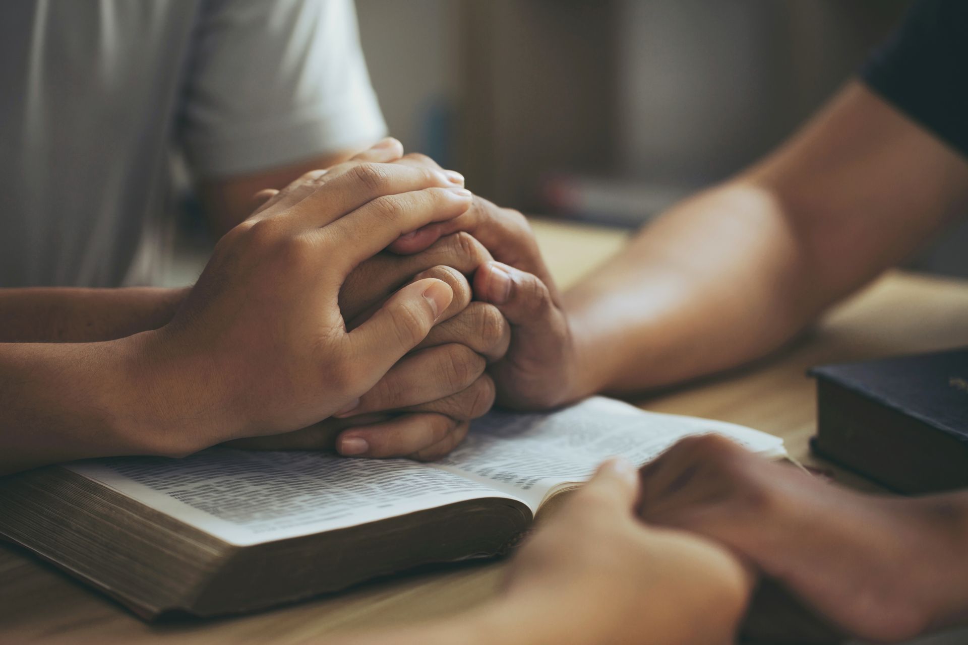 Hands clasped in prayer over an open Bible on a wooden surface.