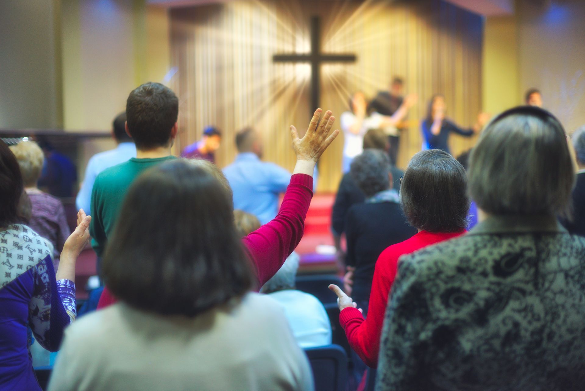 People in a church worship, some with raised hands, facing a stage with a cross and band.