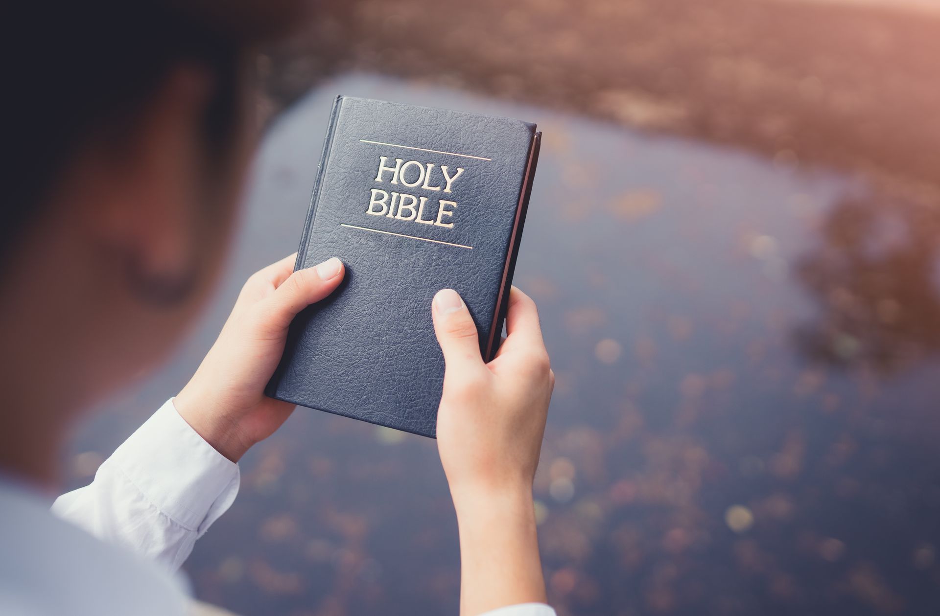 Person holding a dark blue Bible open near water; sunlight.
