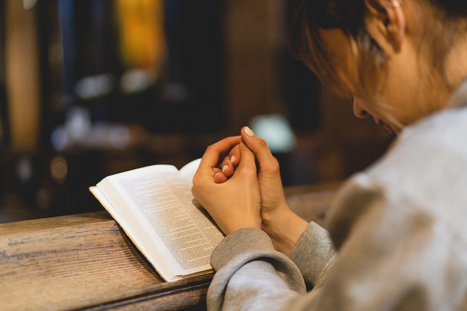 Person with hands clasped in prayer over an open book at a wooden surface indoors.