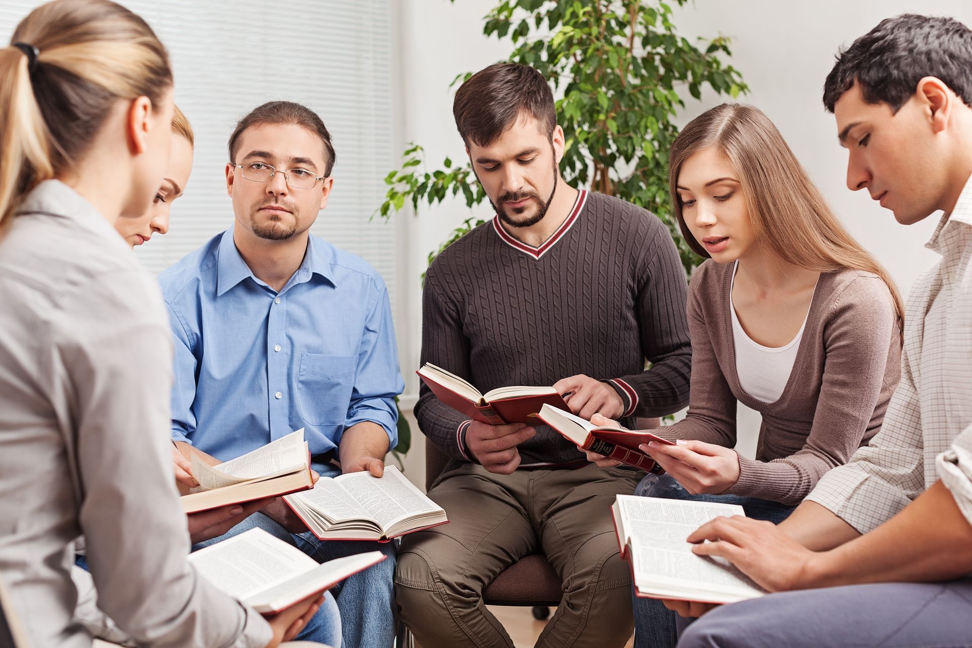 Group of people reading books, seated in a circle indoors, possibly a discussion or study session.