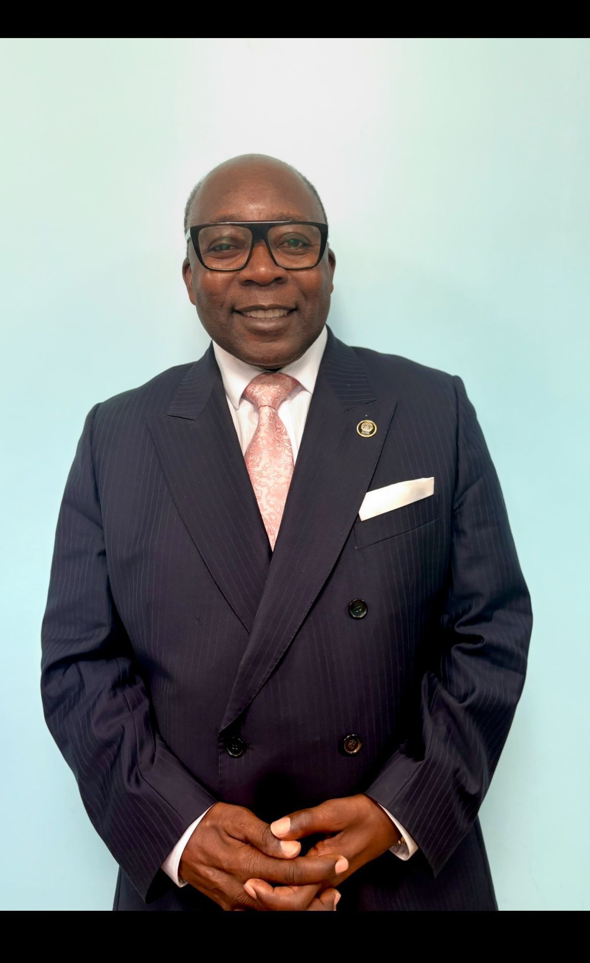 Man in a dark suit, wearing glasses, and a pink tie, smiling, indoors.