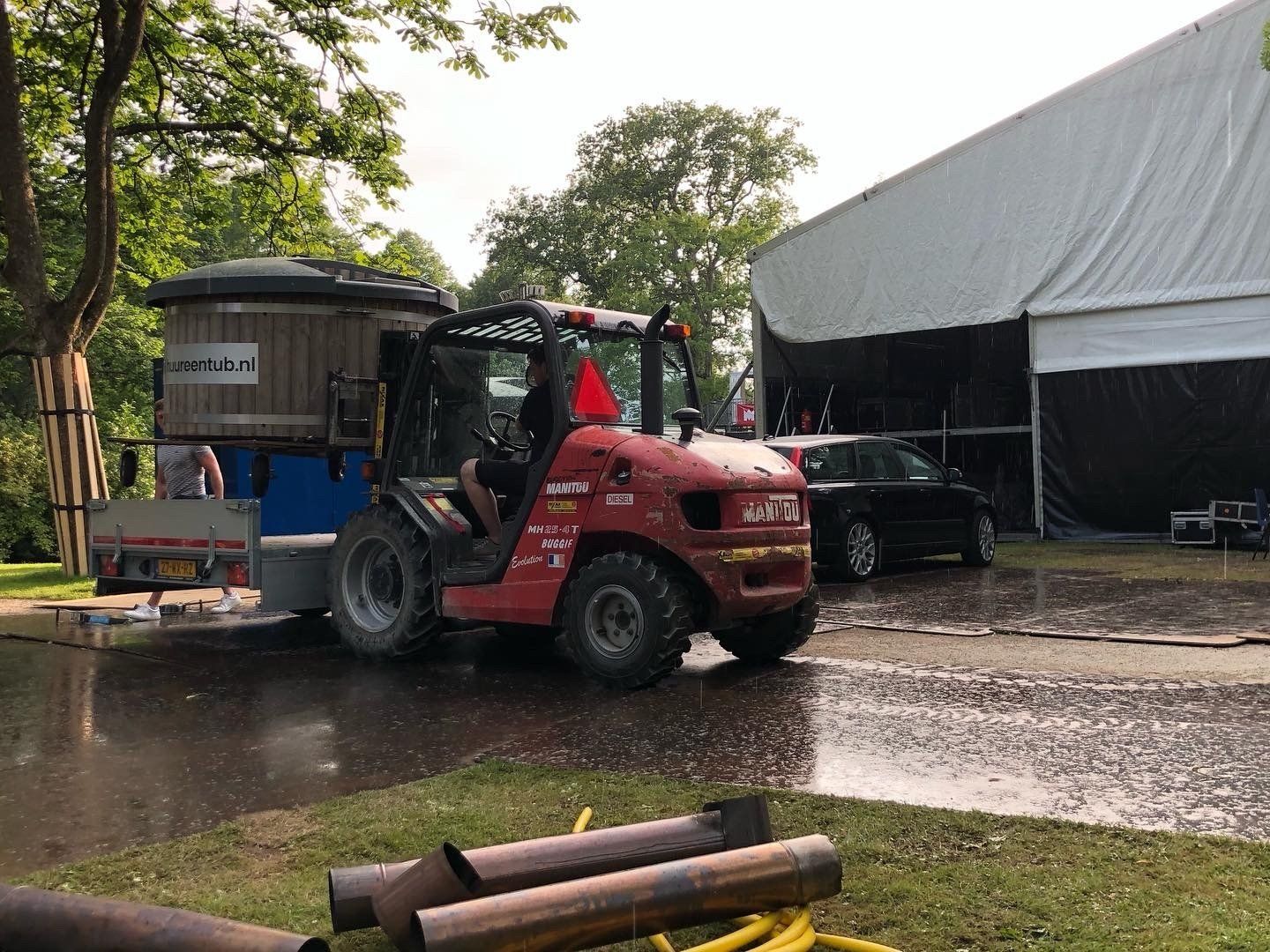 Een rode vorkheftruck tilt een hot tub over een natte weg voor een witte tent.