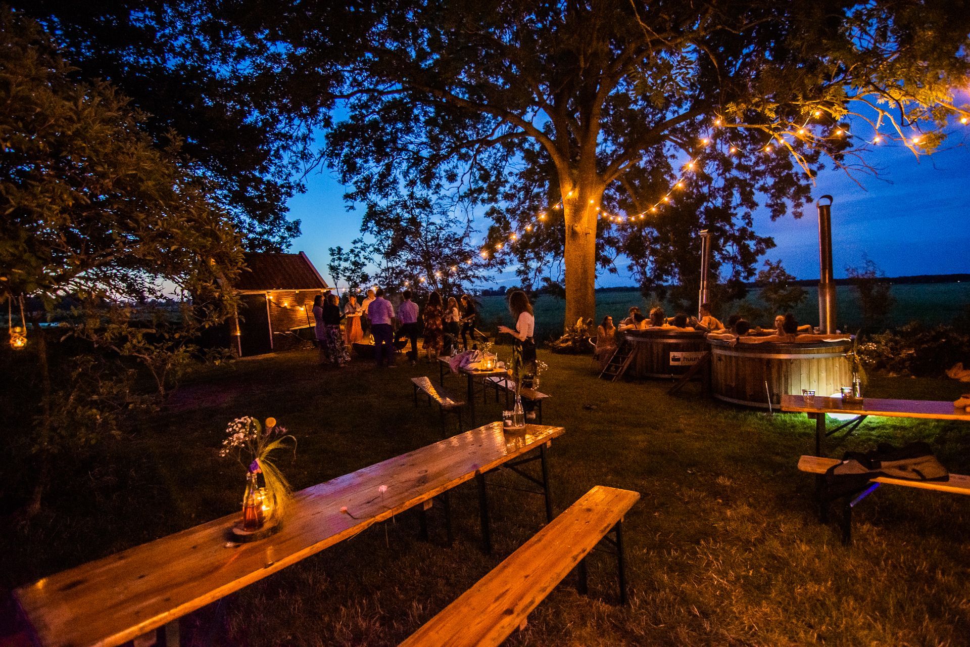 's Avonds zit een groep mensen aan tafels onder een boom met op de achtergrond twee hot tubs. 