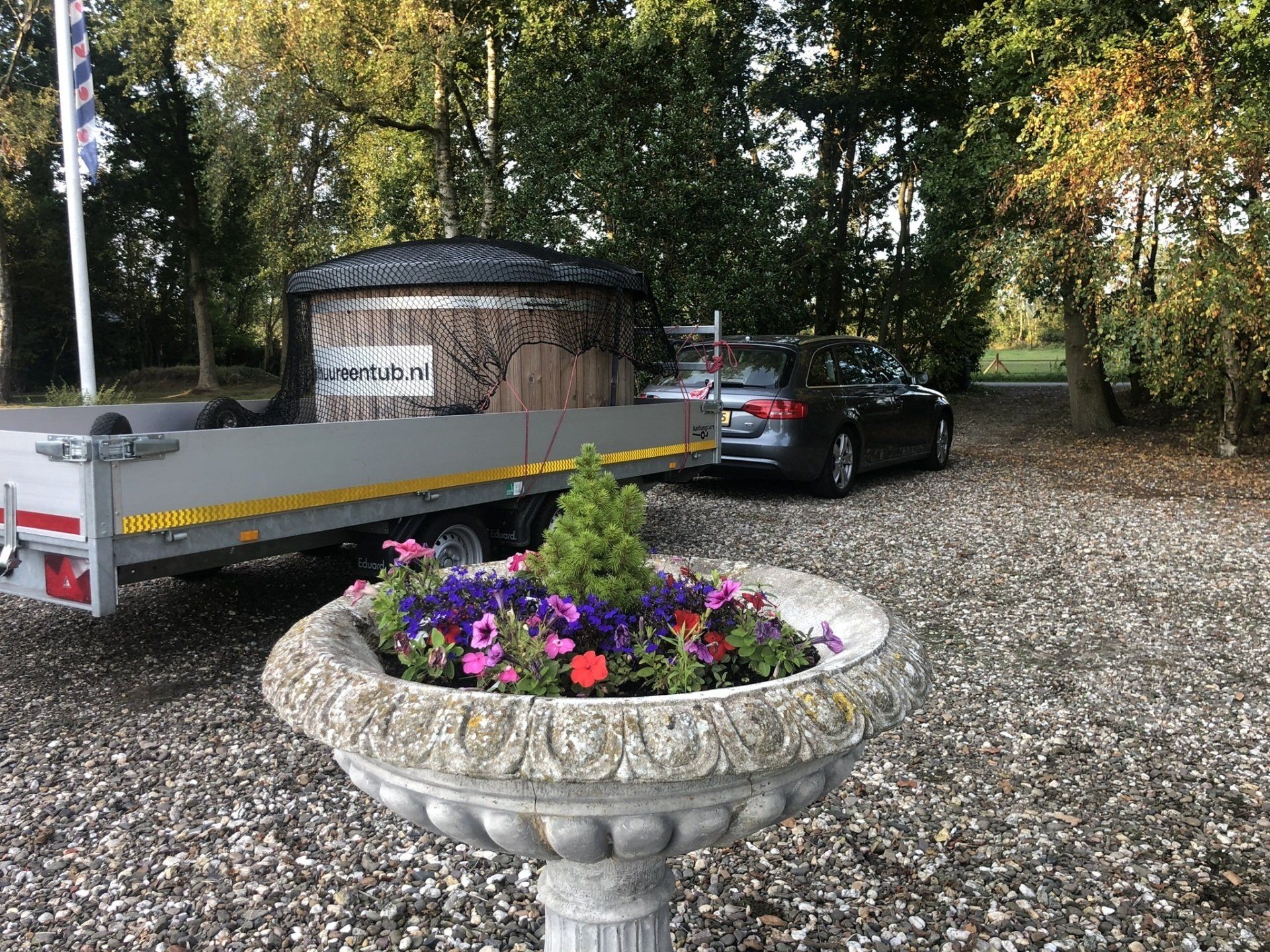 Een auto met aanhanger en hottub staat geparkeerd op een grindpad naast een plantenbak vol bloemen.
