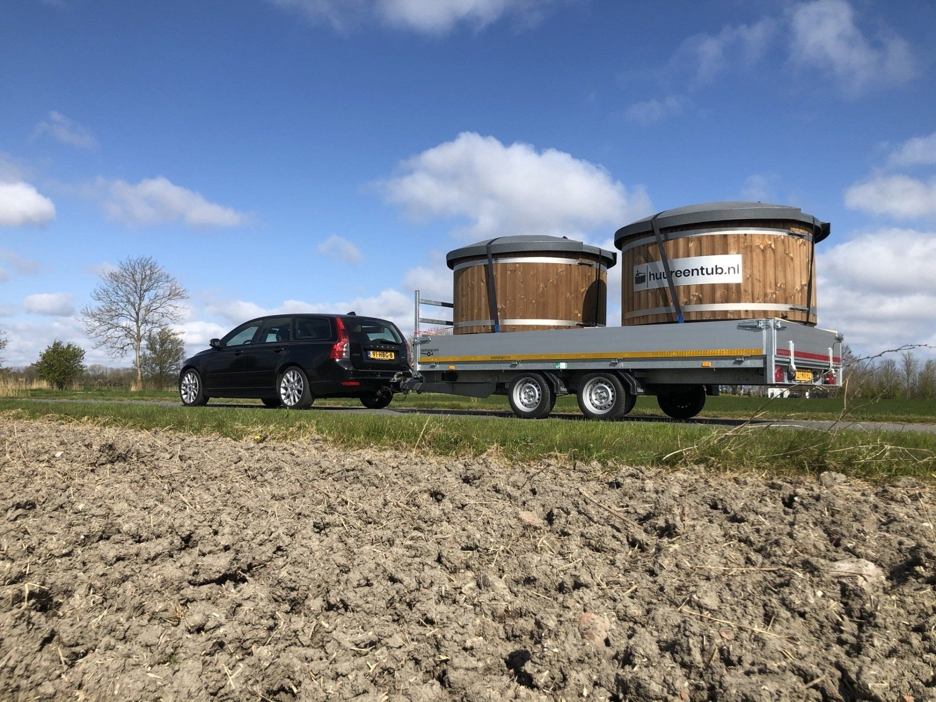 Een auto trekt een aanhanger met daarop twee houten hottubs. 