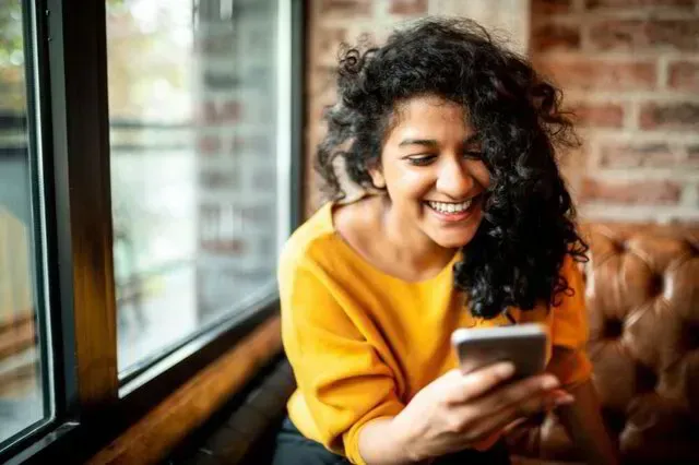 Smiling woman in a mustard sweater using a smartphone by a window in a cozy café