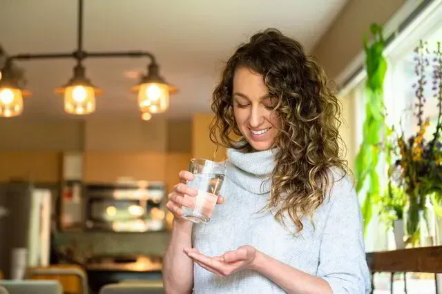 Woman in a bright kitchen holding a glass and looking at her hand, with hanging lights and plants nearby