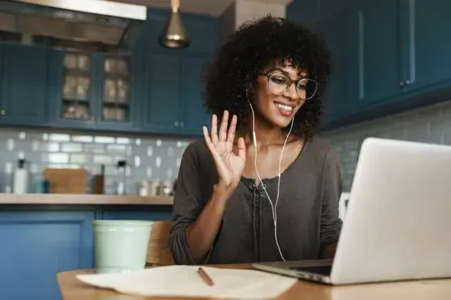 Woman smiling and waving at a laptop in a blue kitchen, with a mug and notebook on the table