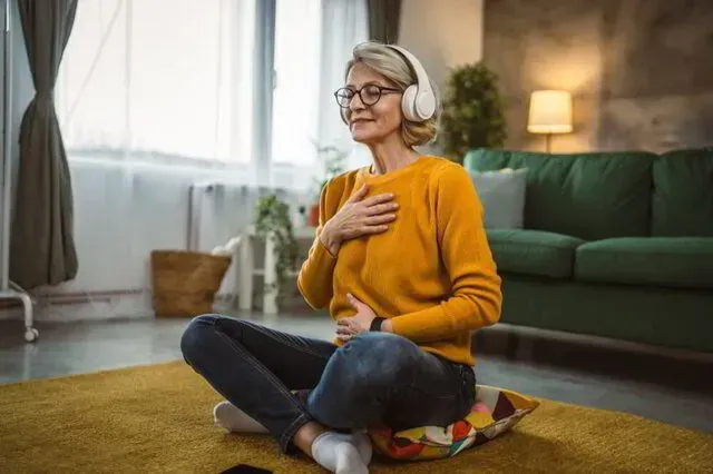 Person meditating on a rug in a living room, wearing orange sweater and white headphones.
