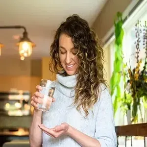 Woman pouring water from a glass into her palm in a bright kitchen with hanging plants