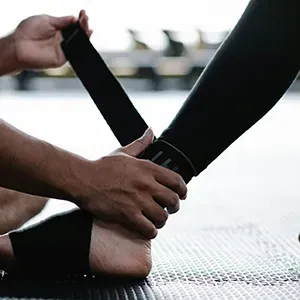 Hands wrapping black ankle straps around a person’s foot on a gym mat