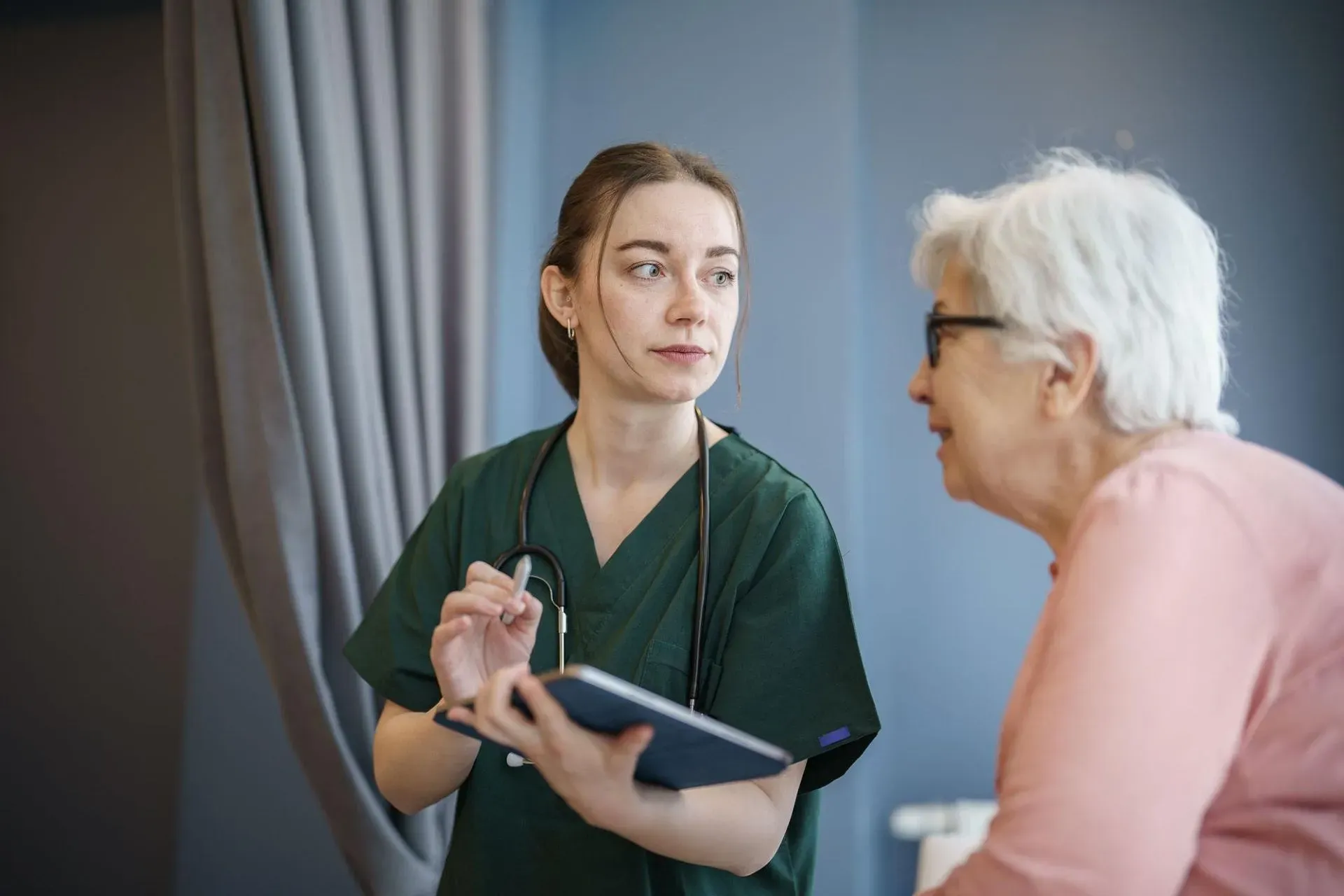 Nurse in green scrubs discussing a tablet with an older woman in a clinic room