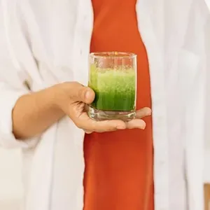 Person holding a small glass of green juice against a white shirt and orange top