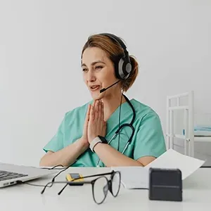 Healthcare worker in teal scrubs with headset, speaking at a desk with laptop and stethoscope