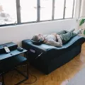 Person reclining on a black couch in a bright room beside a window and side table with electronics