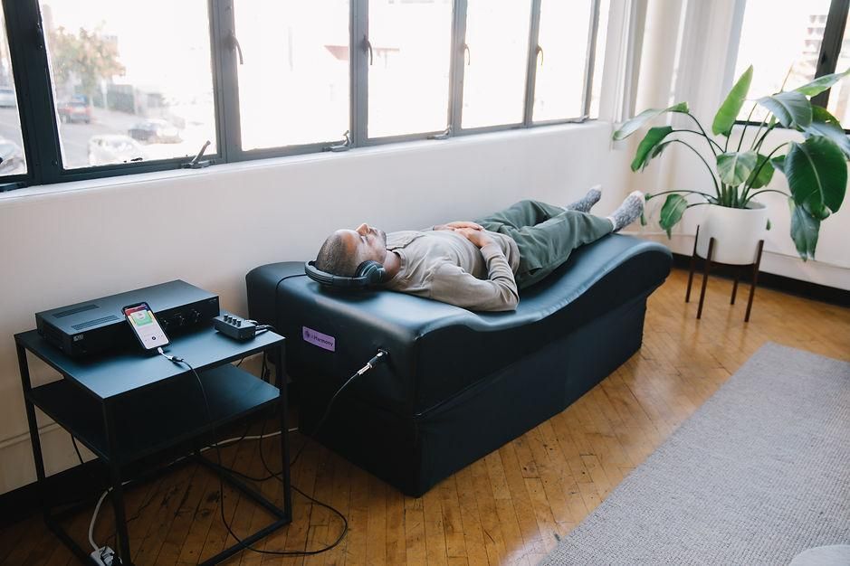 Person lying on a black couch in a bright room with large windows and a small side table.