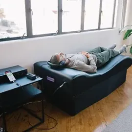 Person lying on a black couch in a bright living room with large windows and indoor plants