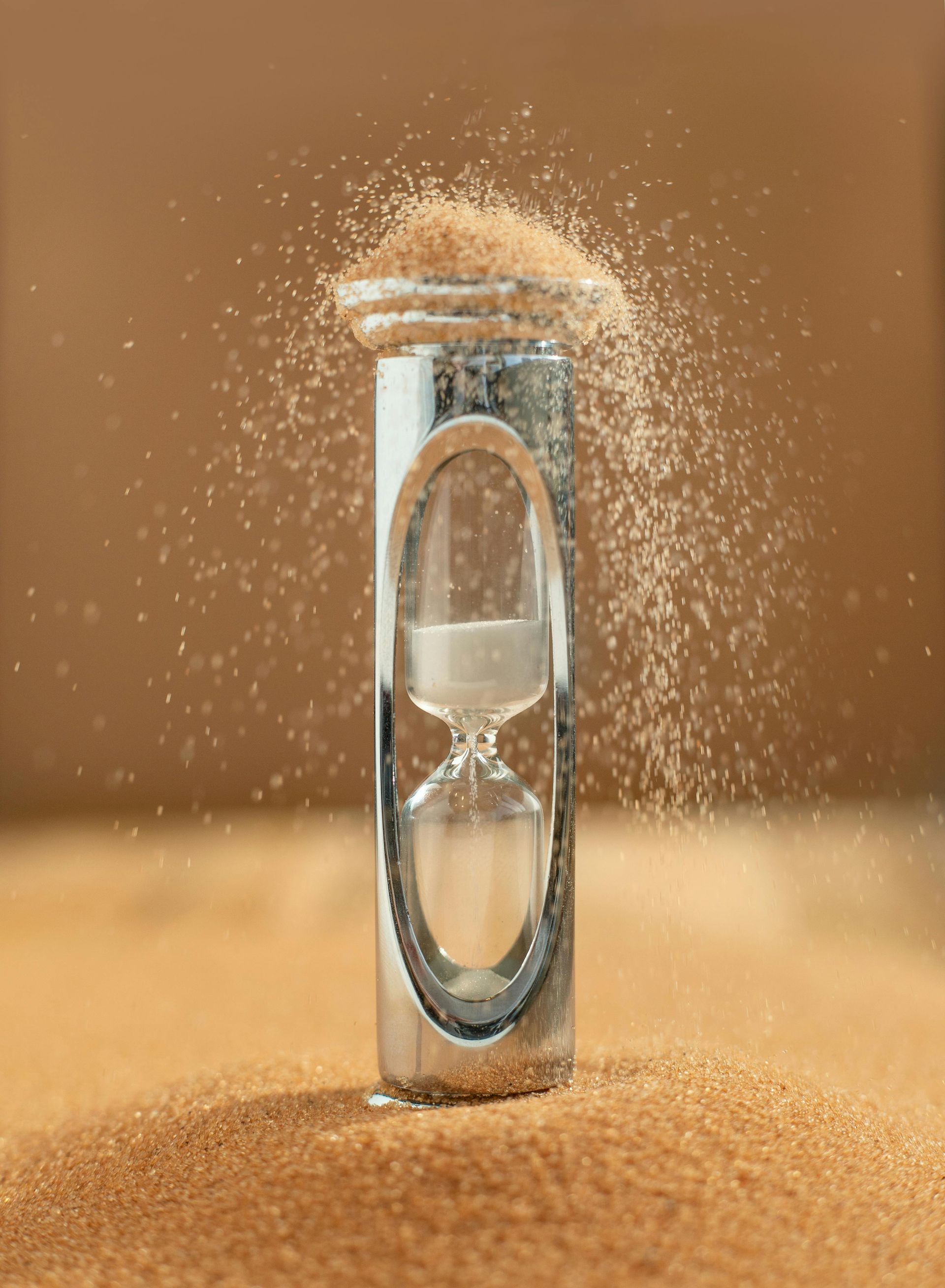Hourglass with sand spilling over the top onto a bed of sand; brown background.