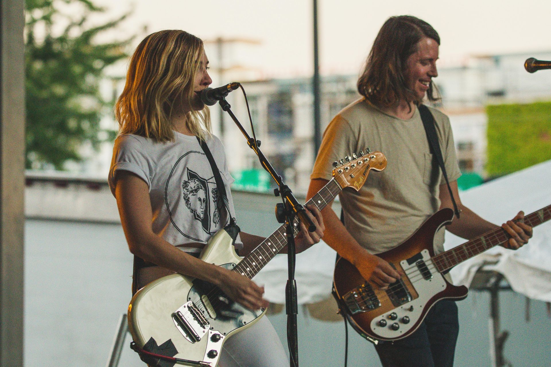 Woman and man playing electric guitars onstage; outdoor setting.