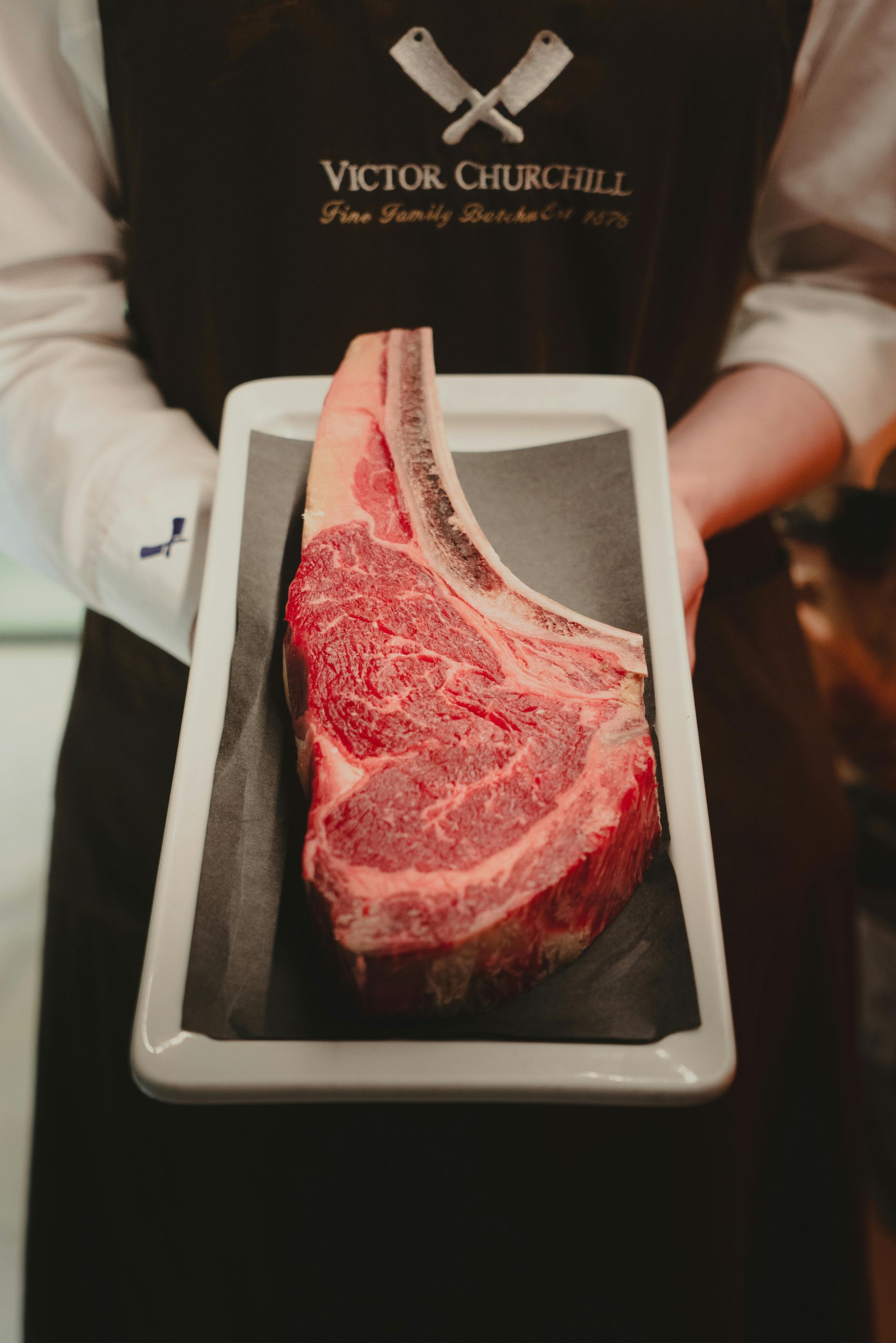 Person holding a large raw ribeye steak on a serving tray, against a restaurant setting.