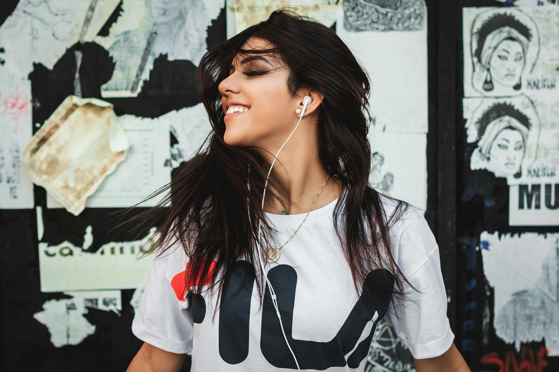Woman smiles, listens to music with earphones, wearing a white FILA t-shirt, in front of a poster-covered wall.