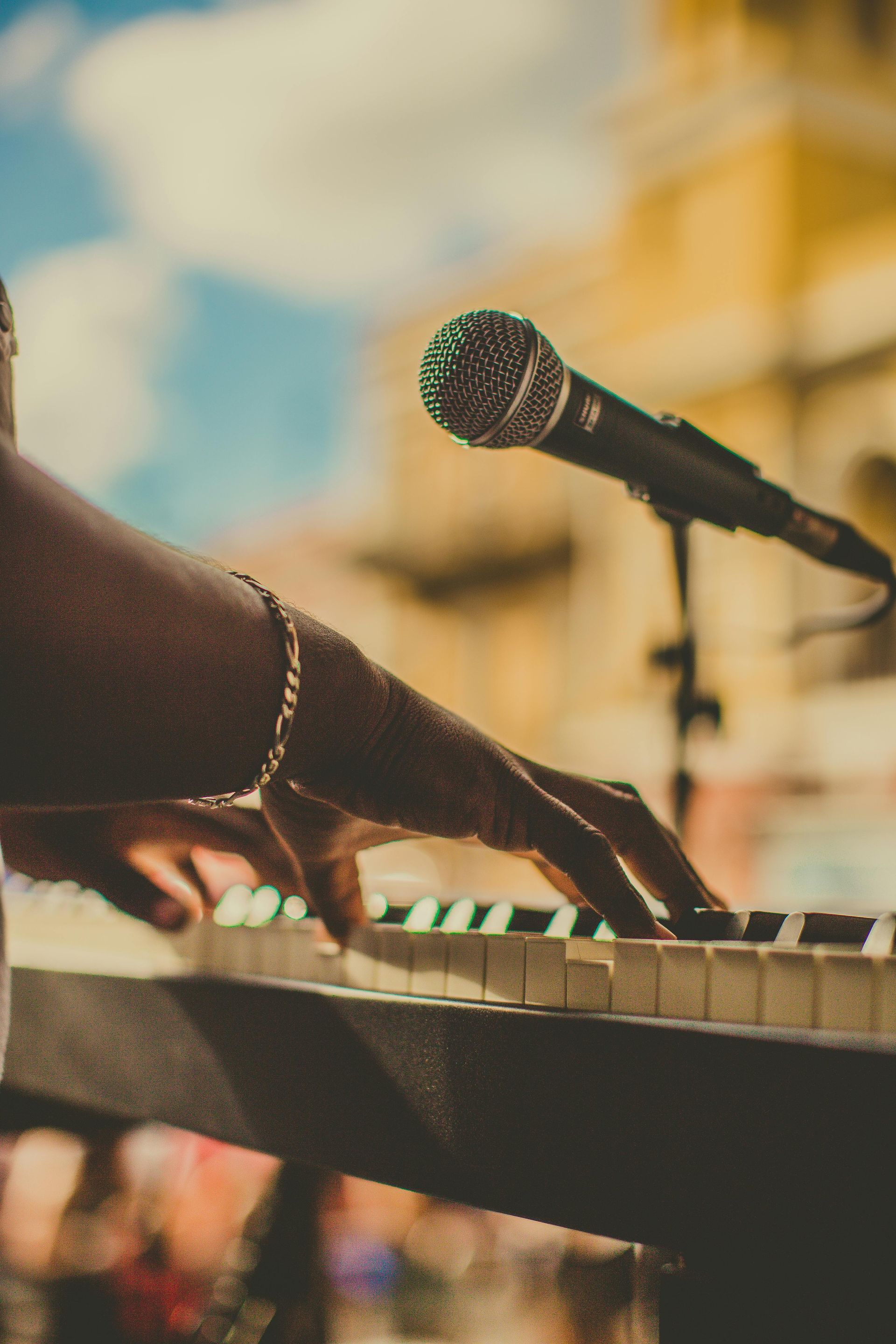 Person's hands playing a keyboard with microphone in front of them, outside in front of a light blue sky.