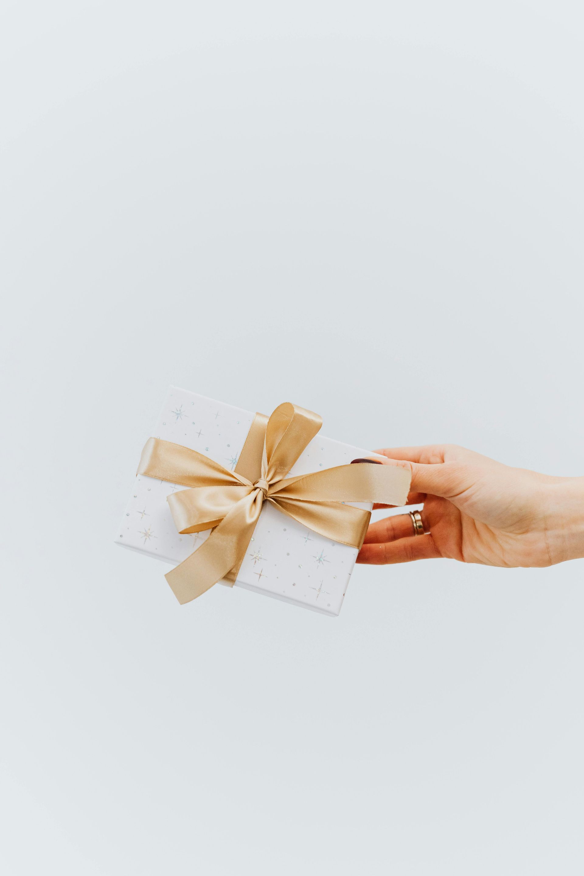 Hand holding a white gift box with a gold ribbon against a light background.