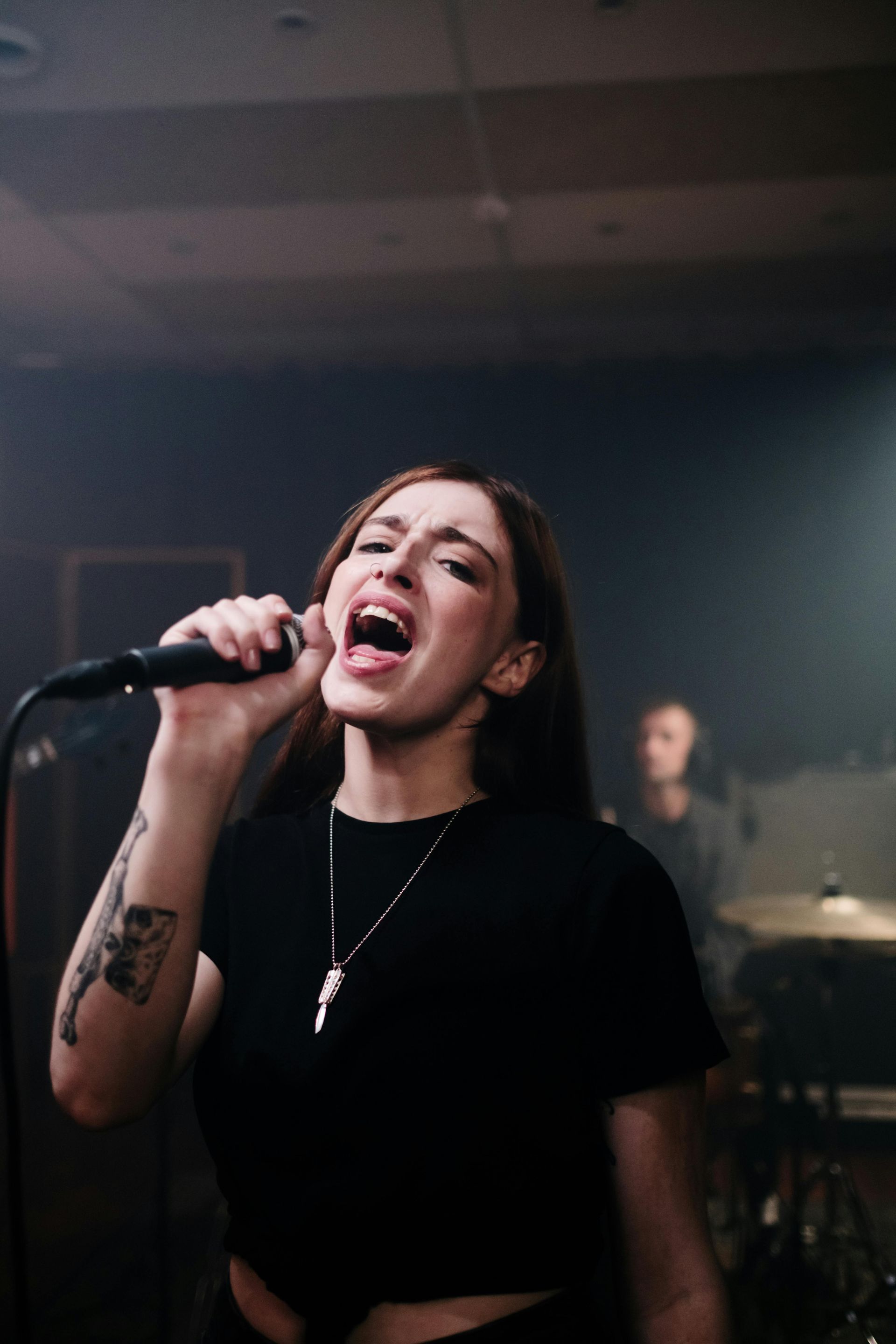 Woman singing into a microphone, intense expression, in a dimly lit performance space.