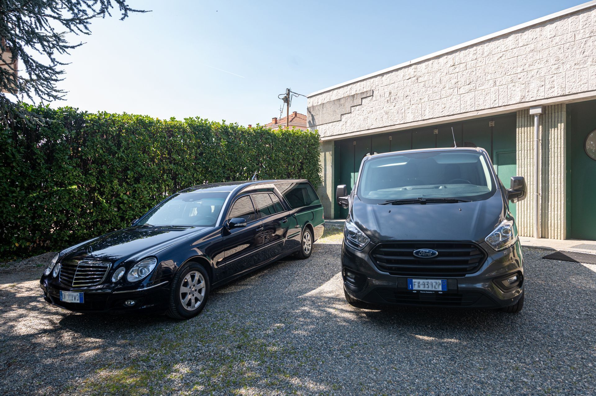 Due auto parcheggiate, una berlina nera e un furgone grigio, in un vialetto di ghiaia accanto a una siepe e a una casa.