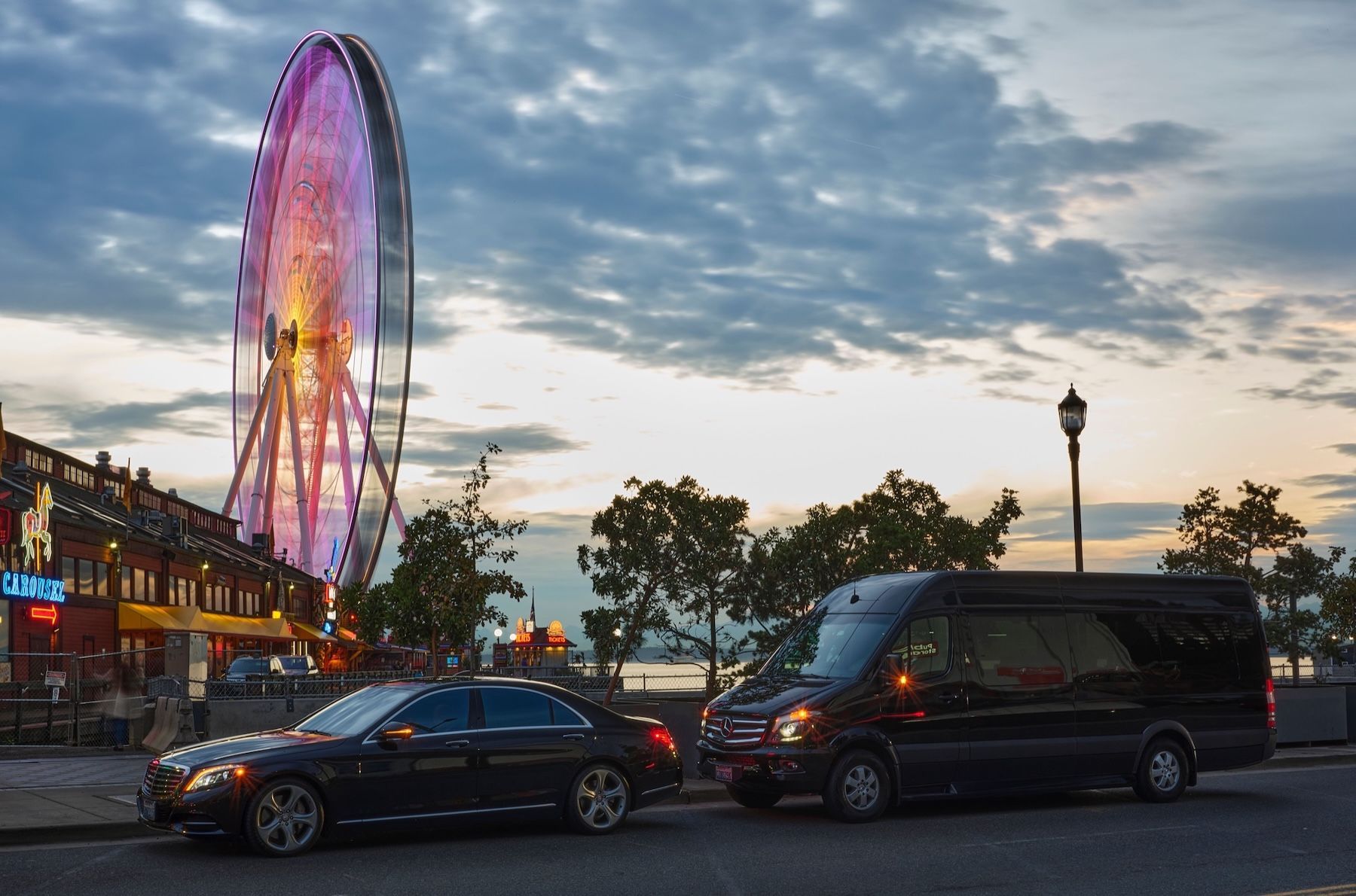 A black car and a passenger van parked on a street near an illuminated Ferris wheel at dusk.