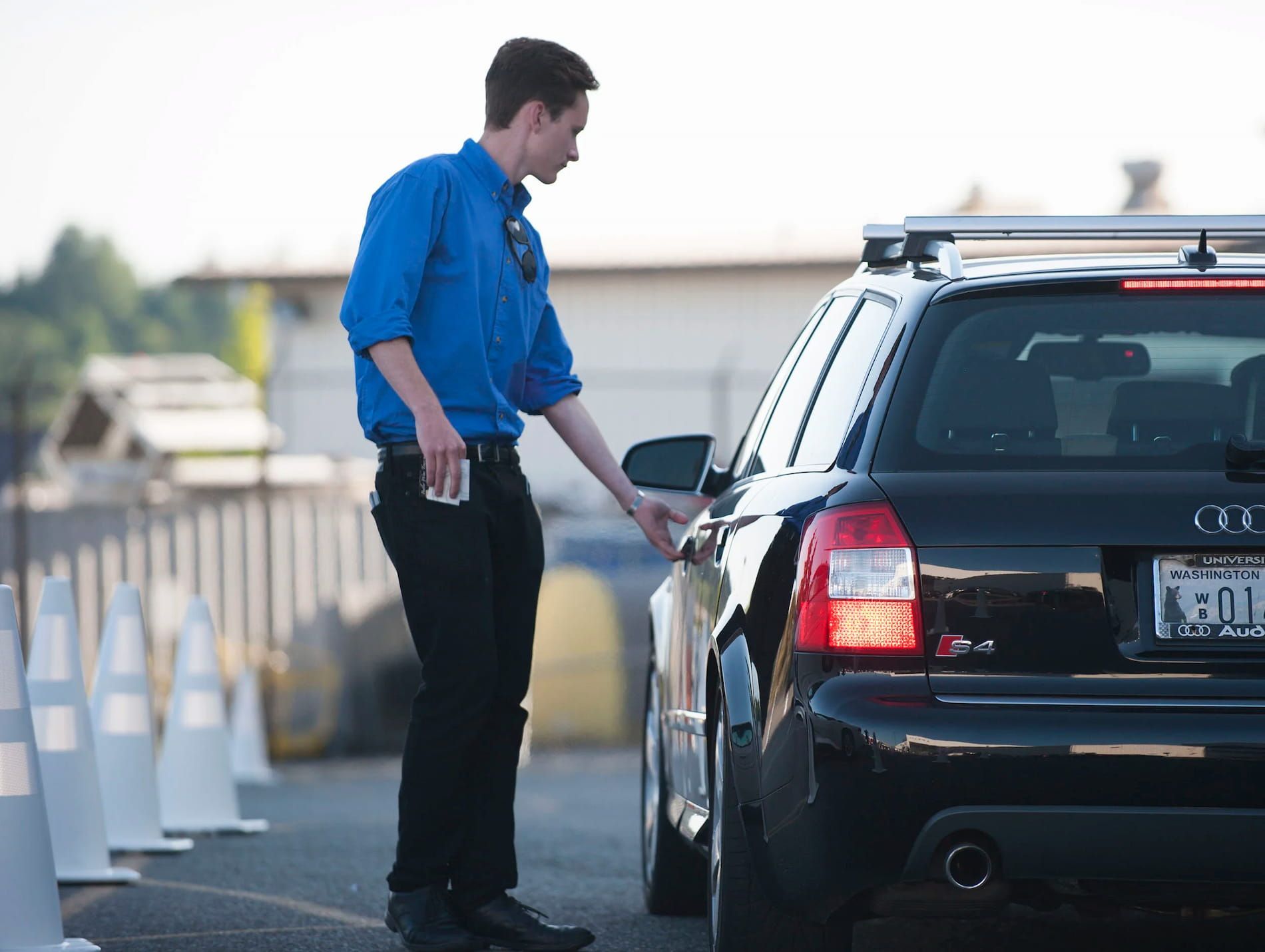 A person in a blue shirt and black pants stands beside a black car, holding the door handle while looking inside.