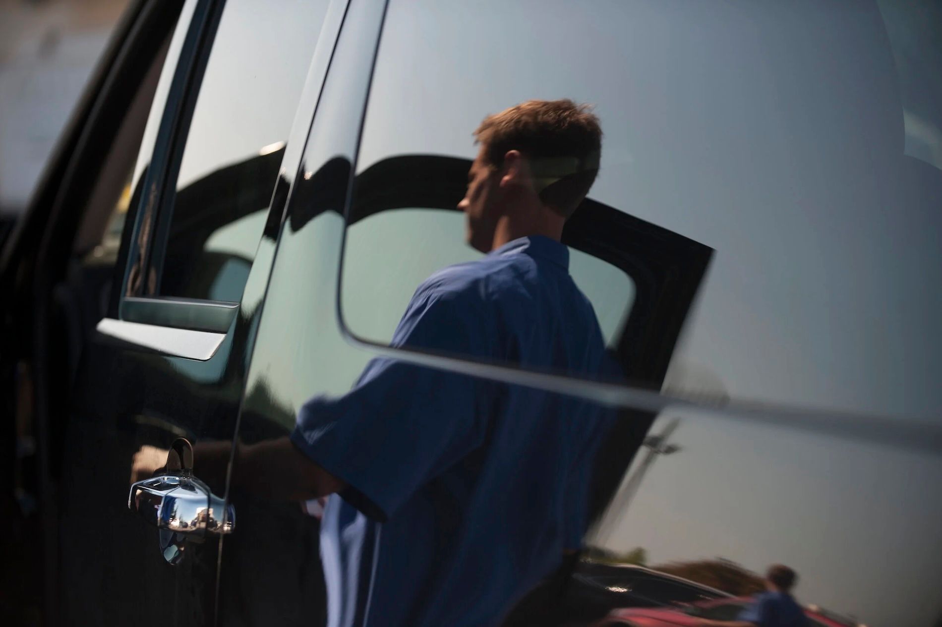 A person in a blue shirt standing by an open car door, seen as a reflection on the vehicle's dark, polished surface.
