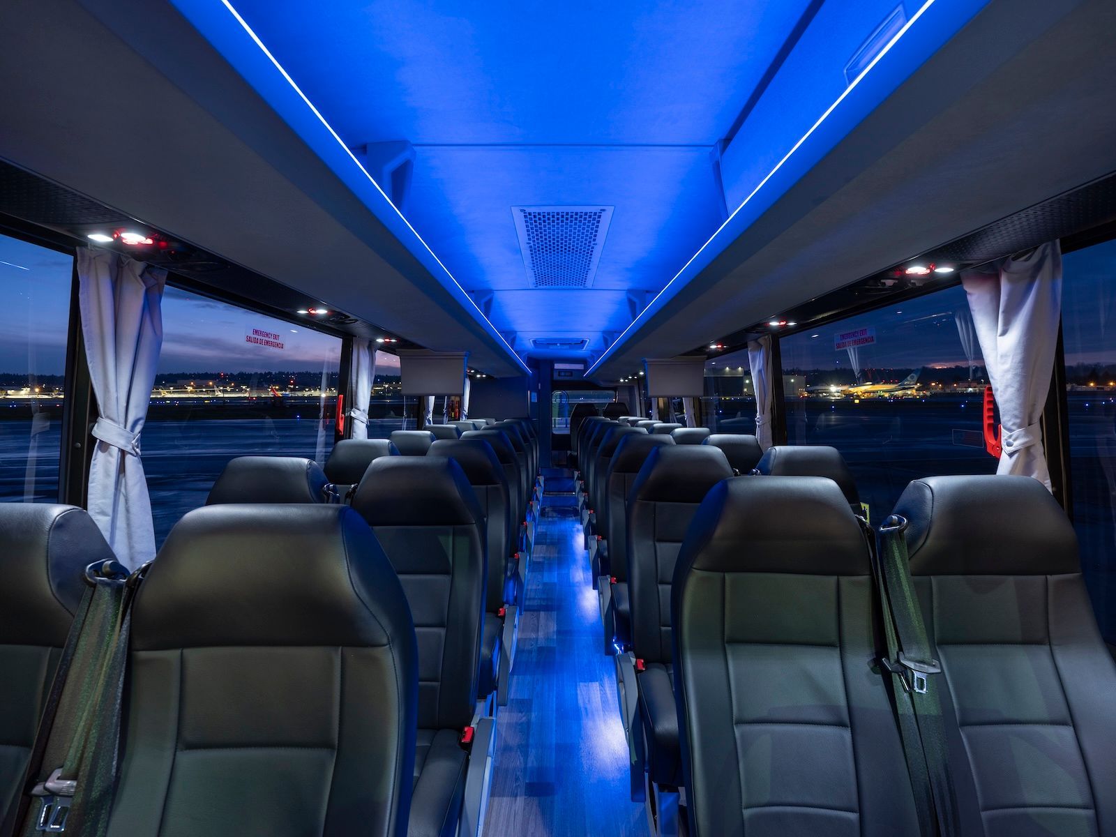 Empty rows of dark leather seats inside a bus with vibrant blue ceiling lights and curtains drawn, facing night windows.
