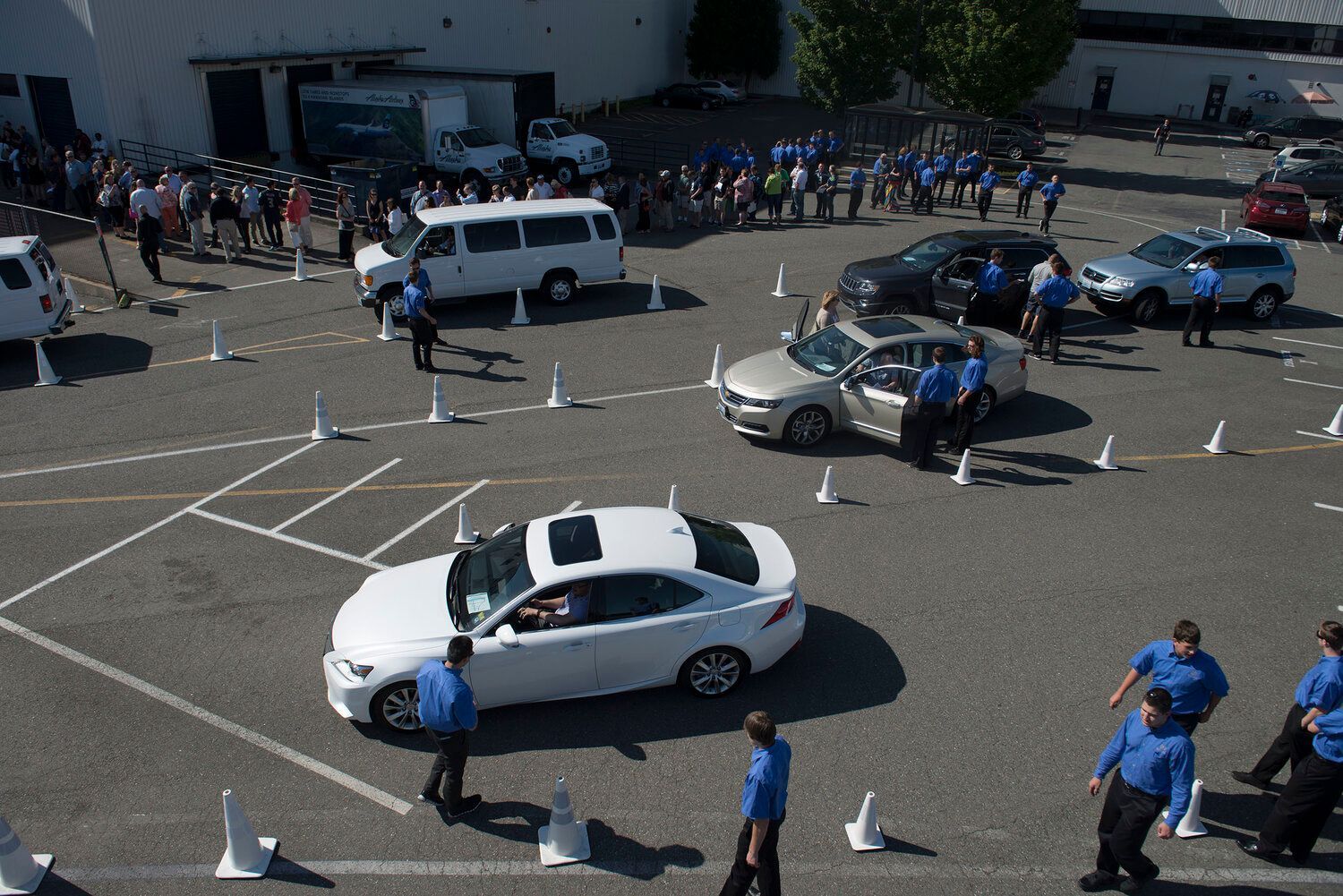 High-angle view of a driving test course with people in blue shirts observing a white car and a tan car navigating cones.
