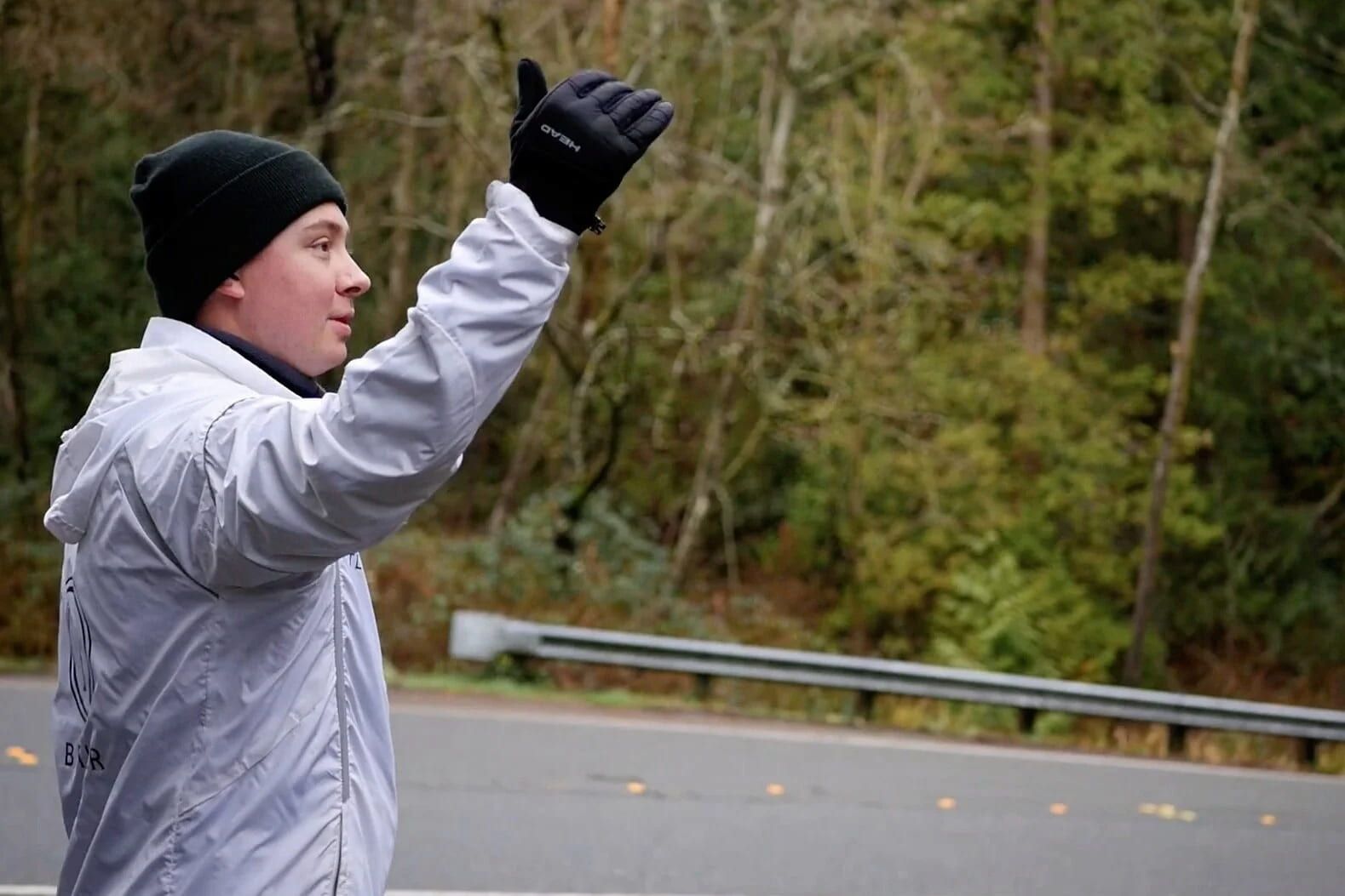 A person wearing a white windbreaker, dark beanie, and gloves stands by a road, signaling with an outstretched hand.