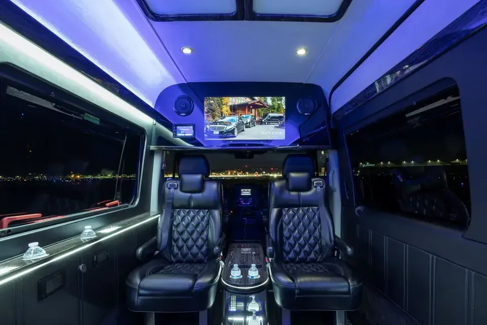 Luxurious interior of a custom van featuring two black leather captain's chairs, blue ambient lighting, and a TV screen.