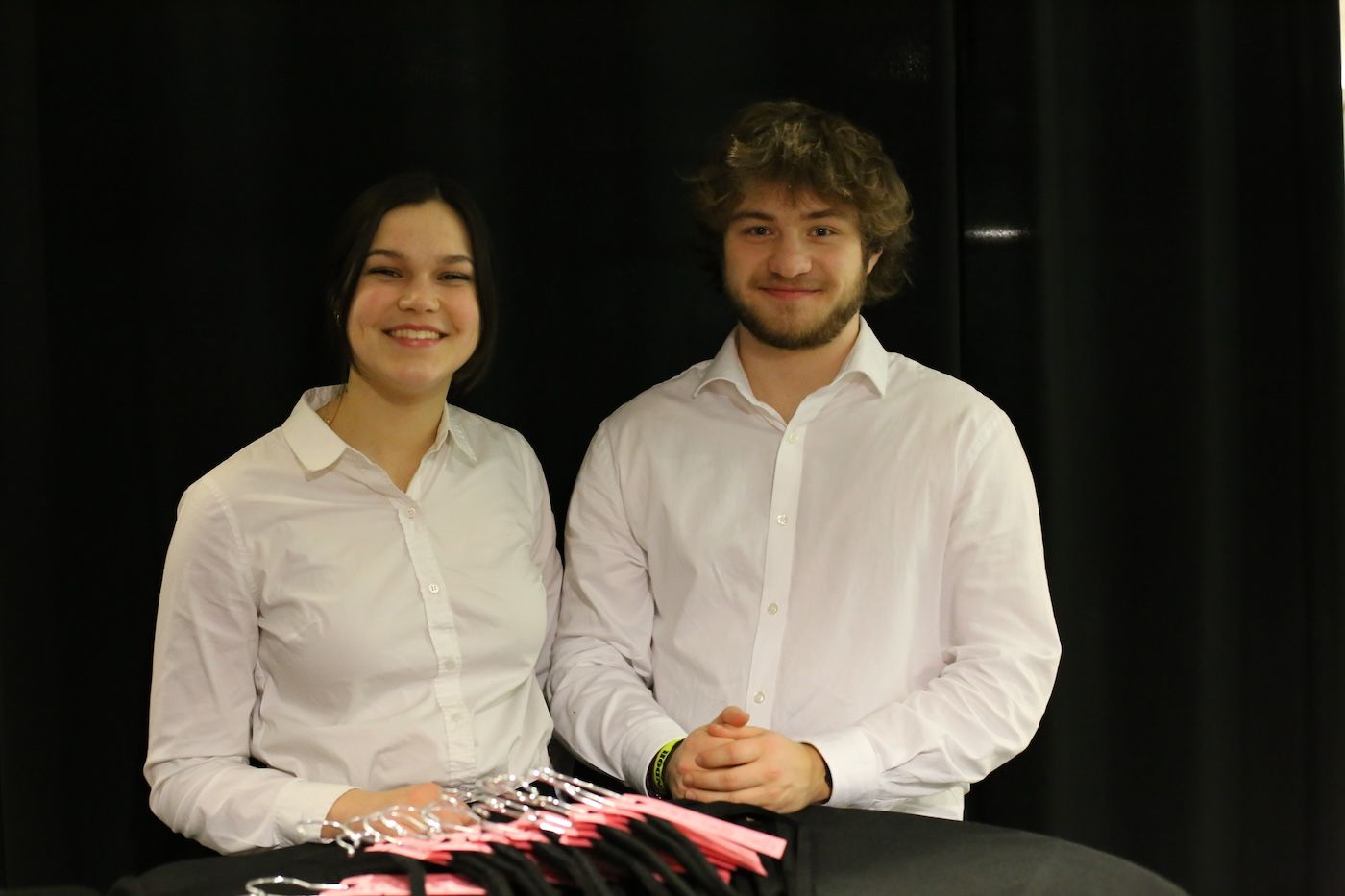 Two people wearing white button-down shirts stand behind a table with pink items against a black backdrop.