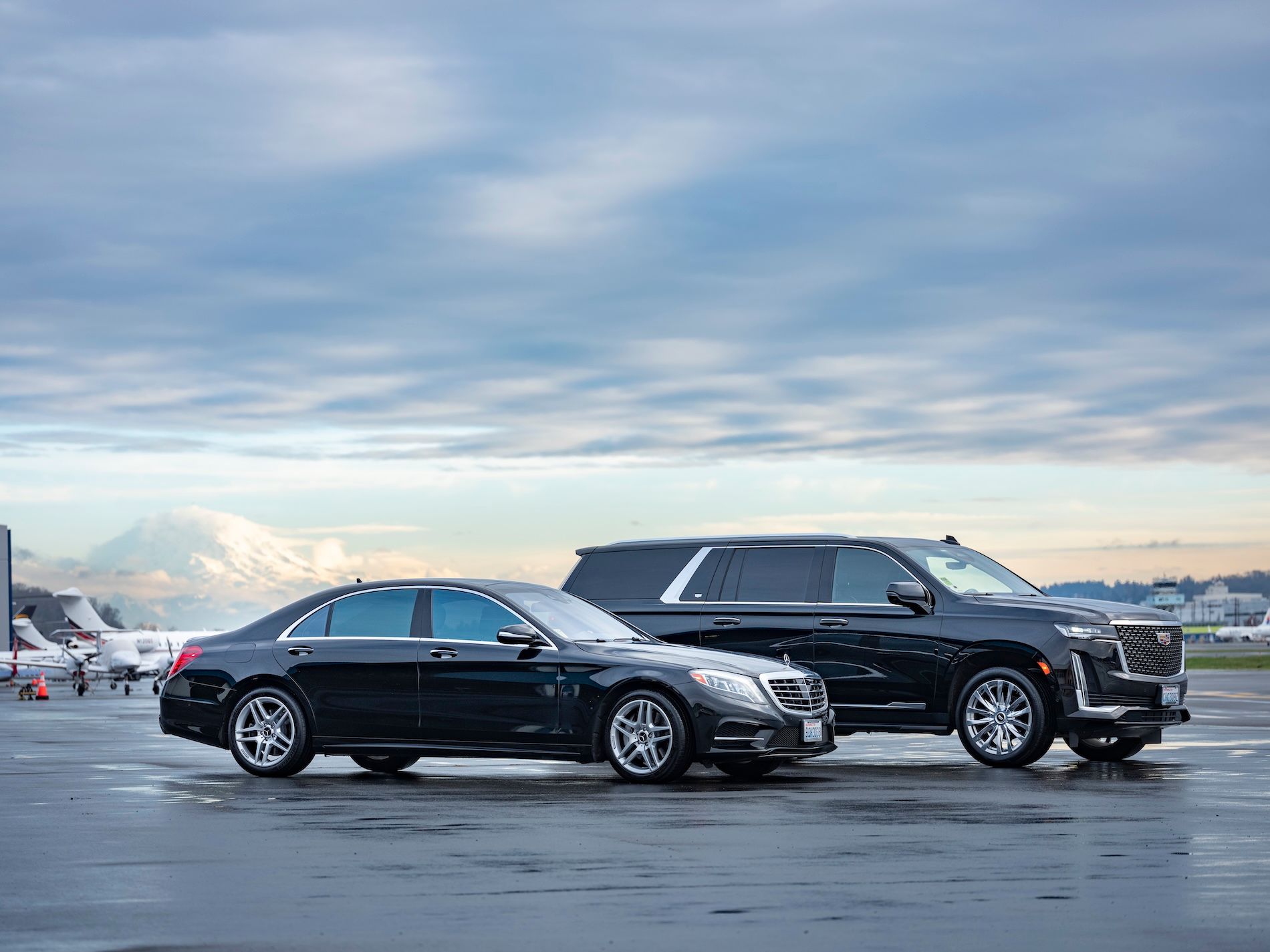 Two black luxury vehicles parked on an airport tarmac with a mountain in the background under a cloudy sky.