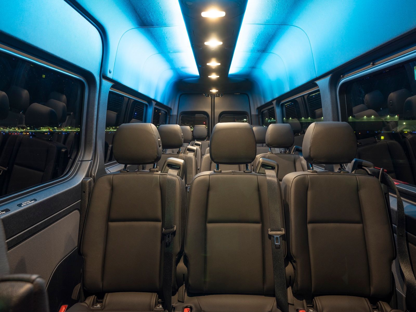 The interior of a passenger van featuring rows of black seats under soft blue ambient ceiling lighting.