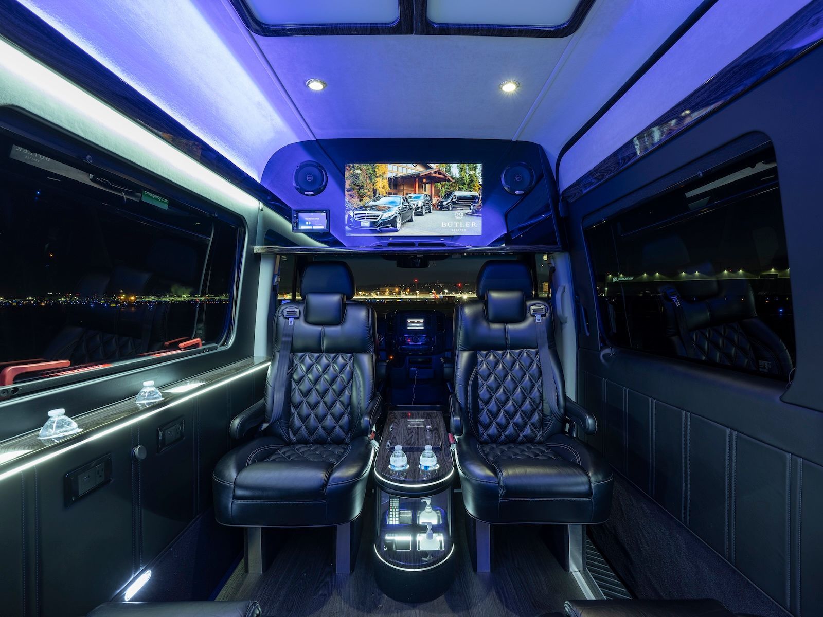 Luxury van interior with black leather seats, blue ambient lighting, and a large TV screen at the front