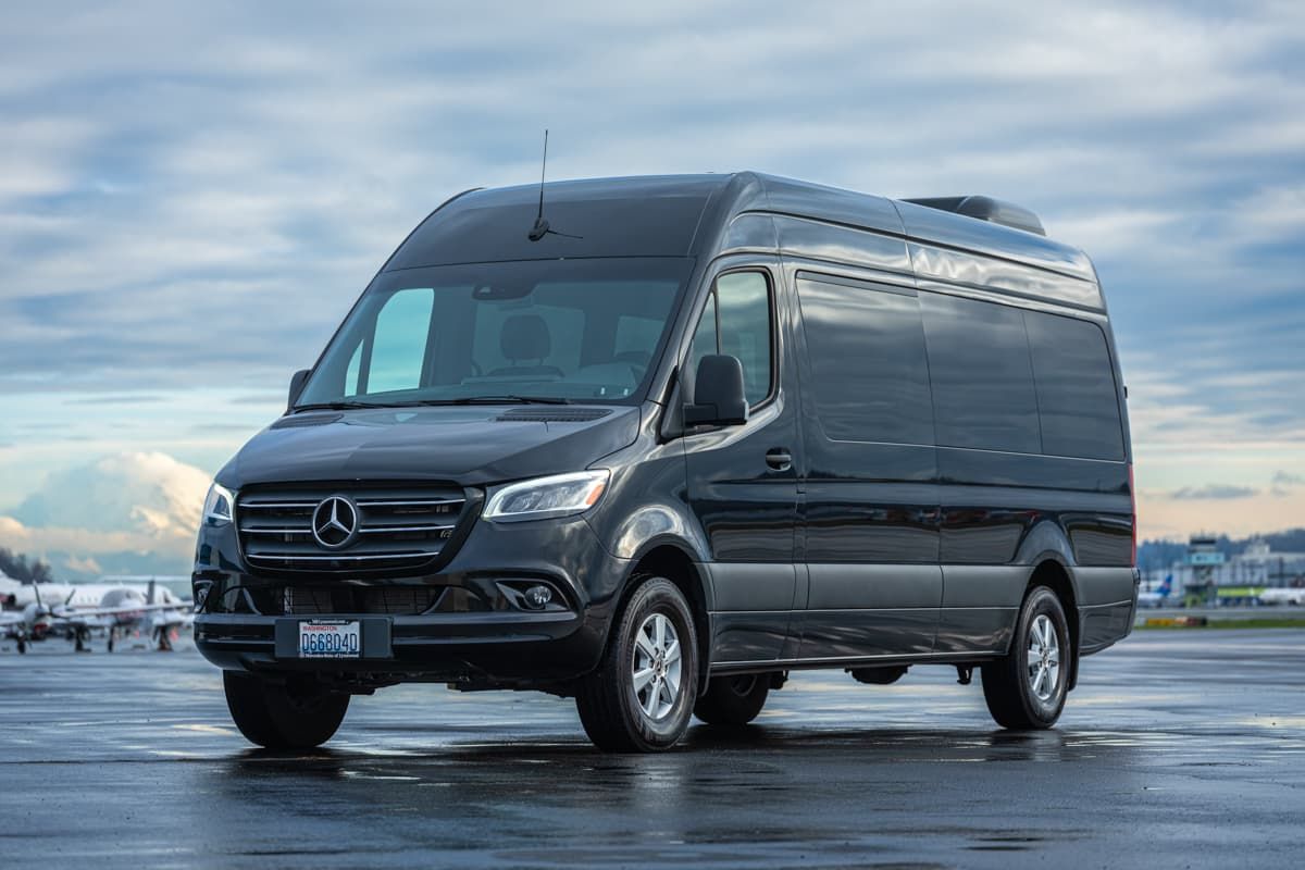 A black Mercedes-Benz Sprinter van parked on an airport tarmac with a cloudy sky in the background.