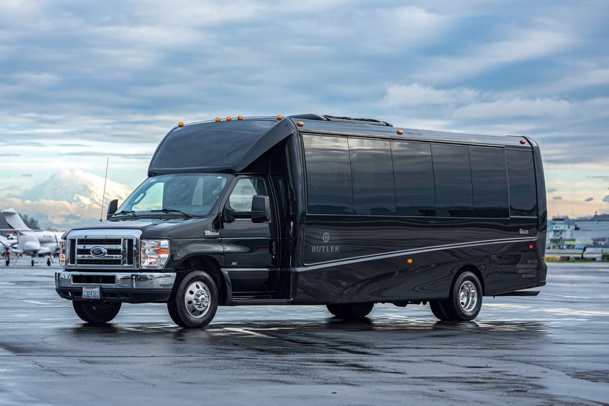 A sleek black passenger shuttle bus parked on a wet tarmac with an airport runway and a distant mountain in the background.