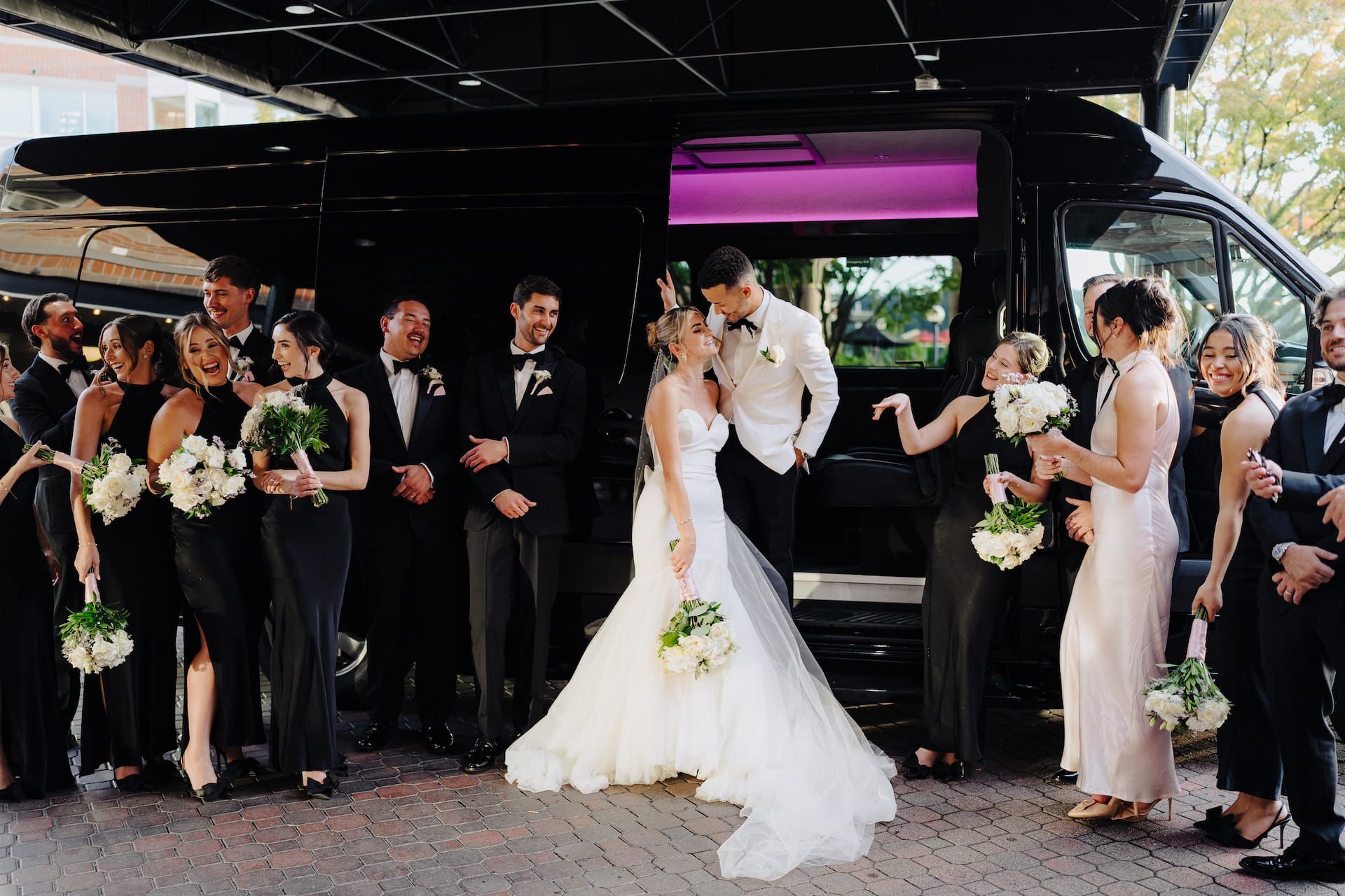 A wedding party in formal attire stands outside a black limo, laughing and smiling while the couple looks at each other.